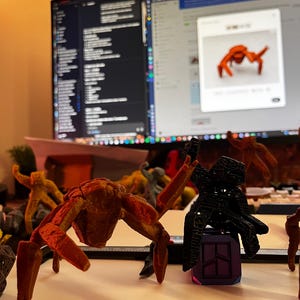 May include: A collection of 3D-printed insect-like sculptures in various colors, including orange, black, and yellow. The sculptures are displayed on a white surface, with a computer monitor in the background.