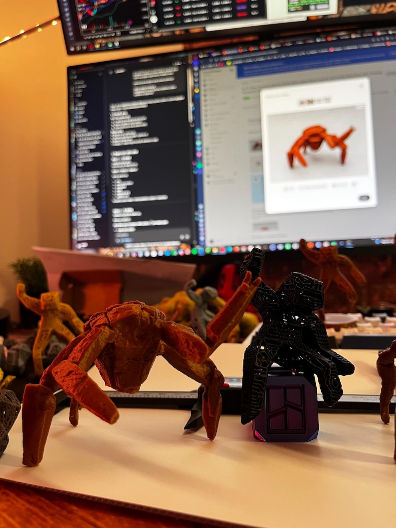 May include: A collection of 3D-printed insect-like sculptures in various colors, including orange, black, and yellow. The sculptures are displayed on a white surface, with a computer monitor in the background.