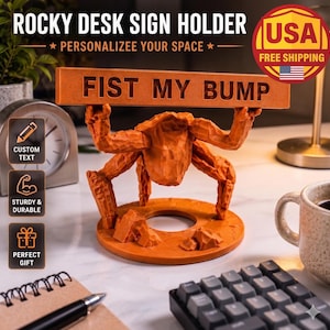 May include: An orange desk sign holder featuring a sculpted figure with multiple limbs holding a horizontal rectangular plaque with the text FIST MY BUMP. The base is circular with rock-like textures, and the figure has a faceted, carved appearance.