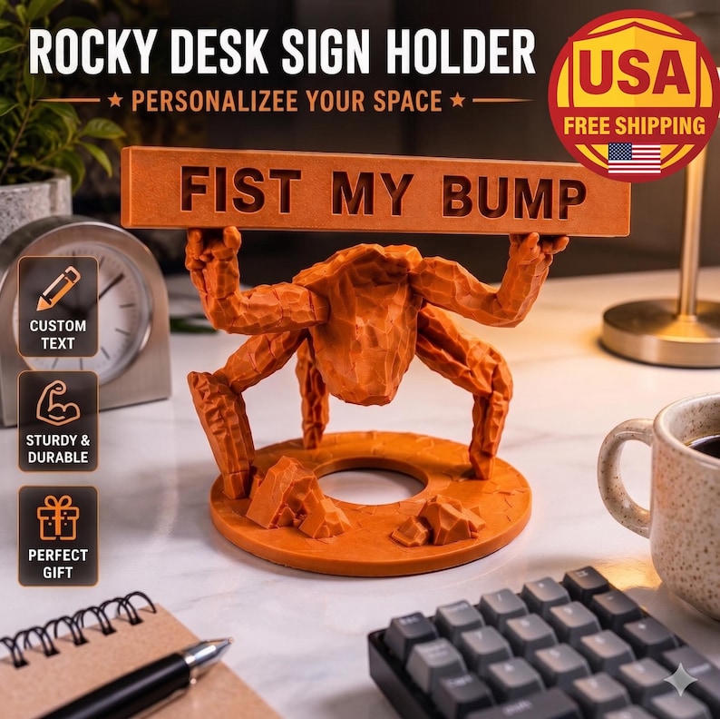 May include: An orange desk sign holder featuring a sculpted figure with multiple limbs holding a horizontal rectangular plaque with the text FIST MY BUMP. The base is circular with rock-like textures, and the figure has a faceted, carved appearance.