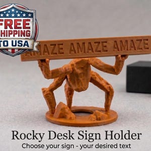 May include: A brown, textured, rock-like figure with four limbs standing on a circular base, holding a horizontal rectangular bar above its head. The bar features the embossed text "AMAZE AMAZE AMAZE" in capital letters across its front surface.