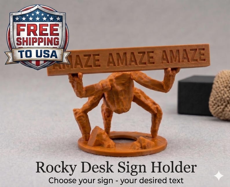 May include: A brown, textured, rock-like figure with four limbs standing on a circular base, holding a horizontal rectangular bar above its head. The bar features the embossed text "AMAZE AMAZE AMAZE" in capital letters across its front surface.