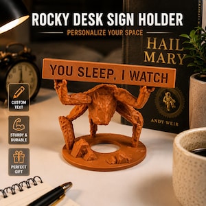 Puede incluir: Un soporte para letreros de escritorio de color marr&oacute;n con textura, que presenta una figura esculpida con m&uacute;ltiples extremidades que sostiene una placa rectangular horizontal. La placa tiene el texto "YOU SLEEP, I WATCH" en may&uacute;sculas.