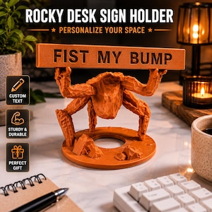Puede incluir: Un soporte de letrero de escritorio naranja con una criatura texturizada similar a una roca con m&uacute;ltiples extremidades que sostienen una barra rectangular horizontal. La barra tiene el texto FIST MY BUMP en relieve. Base circular con detalles.