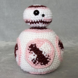 May include: A crocheted plush toy in the shape of a droid with a white, pink, and brown color scheme. The droid has a single black eye and a round body.