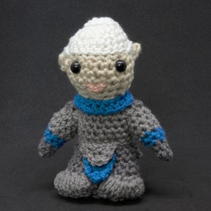 May include: A crocheted toy figure with a gray body, blue scarf, and white hat. The toy has black eyes and a pink mouth.