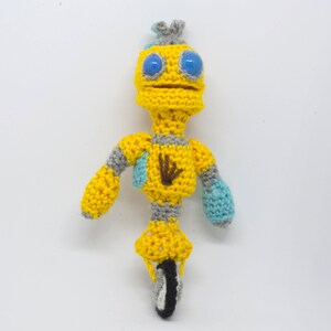 May include: A crocheted yellow robot with blue eyes and a gray body riding a unicycle. The robot has a brown hand on its chest.
