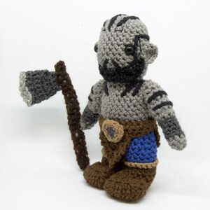 May include: A crocheted grey and brown plush toy with a black beard and a blue and brown belt. The toy is holding a grey and brown crocheted axe.