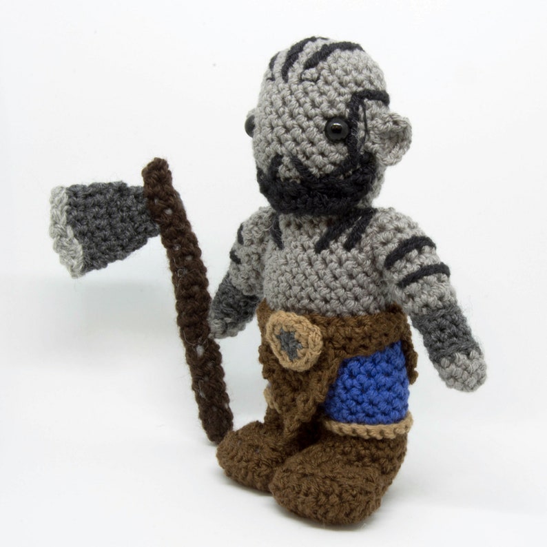 May include: A crocheted grey and brown plush toy with a black beard and a blue and brown belt. The toy is holding a grey and brown crocheted axe.