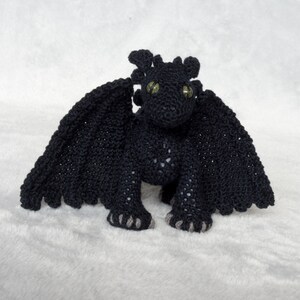Pode incluir: Dragão preto de crochê com olhos verdes e garras cinzas. O dragão está em pé sobre uma superfície branca e fofa.