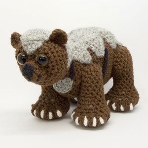 Op de afbeelding: Een gehaakt knuffeldier dat lijkt op een beer met bruin en grijs bont. De beer heeft grote bruine ogen en witte klauwen.