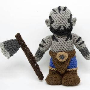 May include: A crocheted grey and black orc figurine holding a grey and brown crocheted axe. The orc is wearing a brown belt with a grey buckle and blue trousers.