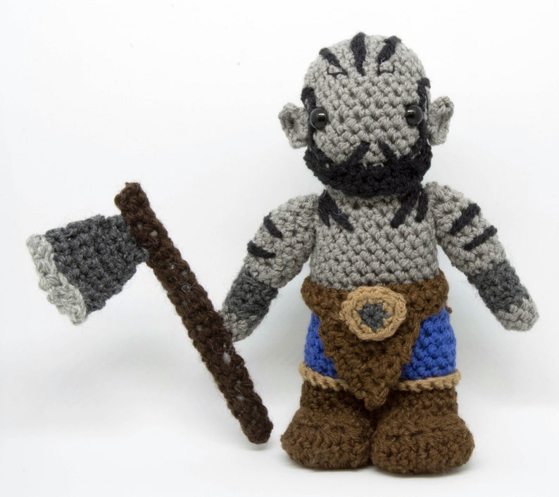 May include: A crocheted grey and black orc figurine holding a grey and brown crocheted axe. The orc is wearing a brown belt with a grey buckle and blue trousers.