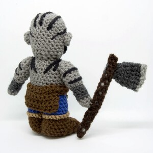 May include: Crocheted grey and brown Orc figurine holding a brown axe. The Orc is wearing blue trousers and brown boots.