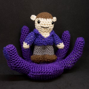 May include: A crocheted purple octopus with a crocheted person in a purple shirt and gray pants sitting on top of its head.