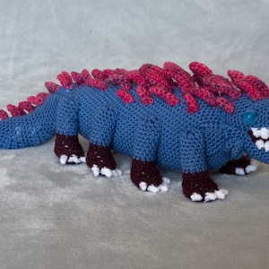 May include: A crocheted blue and pink monster with white teeth and a blue eye. The monster has a spiky pink mane and brown feet.
