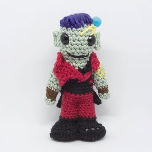 May include: A crocheted green goblin-like figure with purple hair, red and black clothing, and a blue bead on its head.