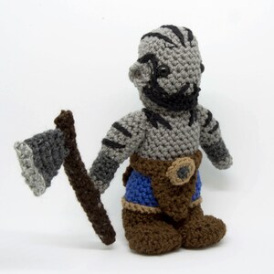 May include: A grey crocheted plush toy of an orc warrior holding a grey and brown crocheted axe. The orc is wearing a brown belt with a round buckle, blue trousers, and brown boots.