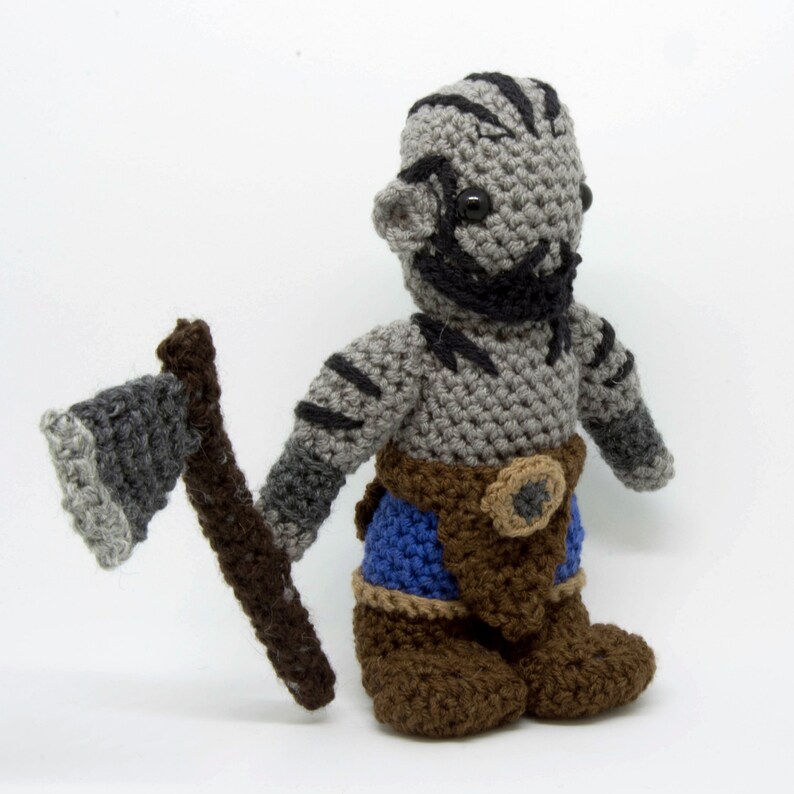 May include: A grey crocheted plush toy of an orc warrior holding a grey and brown crocheted axe. The orc is wearing a brown belt with a round buckle, blue trousers, and brown boots.