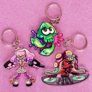 May include: Three colorful keychains featuring characters from the video game Splatoon. The first keychain is a green and purple squid-like creature with a key ring. The second keychain is a pink and white squid-like creature with a key ring. The third keychain is a red and black squid-like creature with a key ring and a DJ turntable.