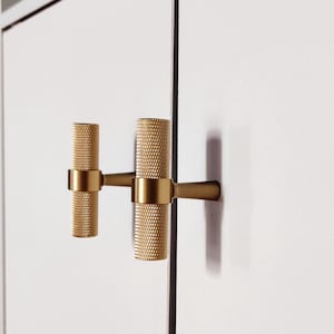 Op de afbeelding: Twee gouden kastknoppen met een getextureerd cilindrisch ontwerp. De knoppen zijn gemonteerd op een witte kastdeur.