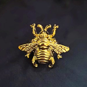 May include: A gold-coloured bee brooch with intricate details. The bee has a textured body and delicate wings.