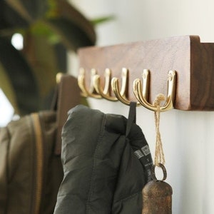 Könnte beinhalten: Ein Wand-Kleiderhaken aus Holz mit sechs goldenen Haken. Zwei Jacken hängen an den Haken, und eine kleine Glocke hängt an einem der Haken.