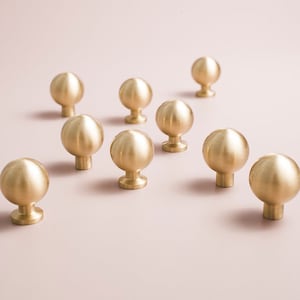 May include: Ten round, polished brass cabinet knobs with a smooth, slightly domed surface. The knobs are arranged in a staggered pattern on a light pink background.