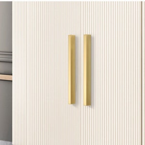 May include: Two gold cabinet pulls with a rectangular shape are mounted on a white cabinet door with vertical grooves.