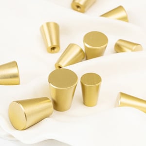 May include: A collection of gold-toned cabinet knobs with a variety of shapes and sizes. The knobs are arranged on a white fabric surface.