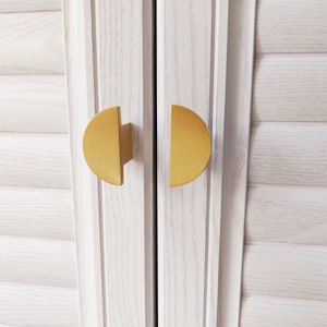 以下が含まれることがあります： 白いキャビネットのドアに、金色の半円形のキャビネットドアノブが2つ付いている。