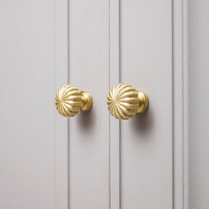 May include: Two gold cabinet knobs with a fluted design, resembling sunflowers, are fixed to a light grey cabinet door. The knobs are round with a domed shape and a polished finish. The cabinet door features vertical paneling.