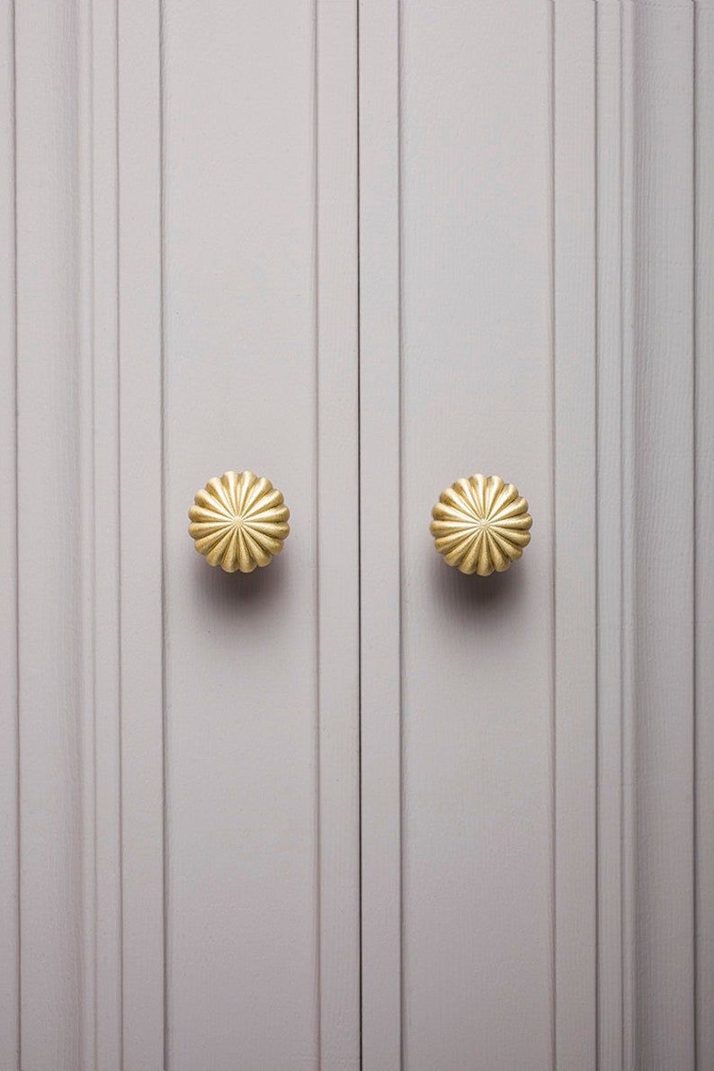 May include: A pair of gold-coloured, flower-shaped knobs on a light grey cabinet door. The door has vertical grooves. The knobs are centred on the door panels and have a ridged design.