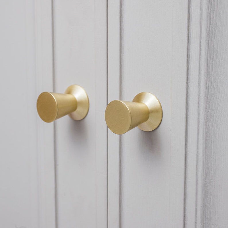 Op de afbeelding: Twee gouden kastknoppen met een cilindrische vorm en een licht taps toelopend ontwerp. De knoppen zijn gemonteerd op een lichtgrijs oppervlak.