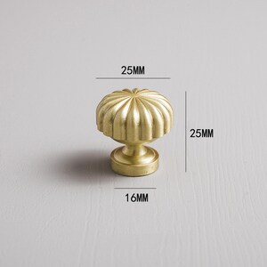 May include: A gold-coloured cabinet knob with a ribbed, rounded top and a pedestal base. The knob measures 2.5cm in height and width, and 1.6cm in diameter at the base. The hardware is designed for furniture and home decor.