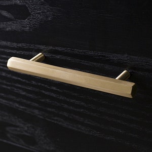 Könnte beinhalten: Nahaufnahme eines goldfarbenen, geometrischen Schubladengriffs, der auf einer schwarzen Holzoberfläche montiert ist. Der Schubladengriff hat eine rechteckige Form mit abgewinkelten Seiten und ist mit zwei kleinen Pfosten am Holz befestigt.