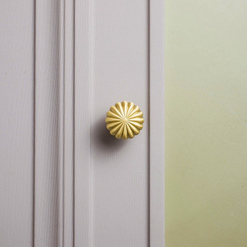 May include: A gold-coloured, flower-shaped cabinet knob is mounted on a light grey, vertically panelled surface. The knob has a detailed, ridged design. The background is a pale green colour.