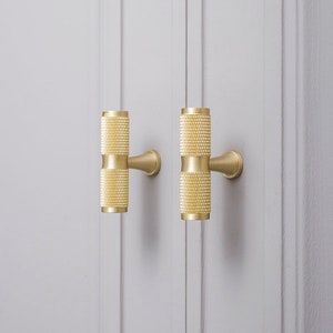 May include: Two gold cabinet knobs with a textured cylindrical design. The knobs are mounted on a white wall.