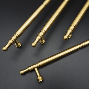 May include: Close-up of several brass-coloured cabinet pulls. The pulls have a cylindrical bar with decorative detailing and curved end pieces. The hardware is arranged diagonally against a dark background, showcasing their design and finish.