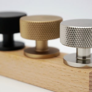Peut inclure: Trois boutons de porte avec une finition texturée. Les boutons sont noirs, dorés et argentés. Les boutons sont présentés sur une base en bois.