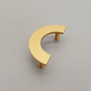 May include: A gold-toned half-circle cabinet pull with rounded edges and a smooth finish.