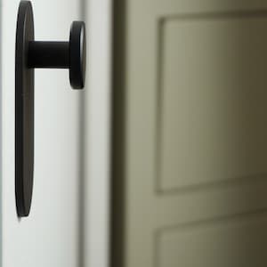 Pode incluir: Um gancho de metal preto montado em uma parede branca. O gancho tem formato oval com uma maçaneta redonda na ponta.