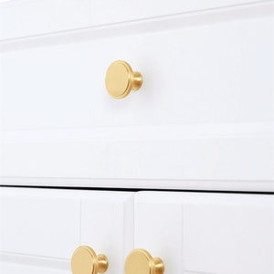 May include: Close-up of white cabinet doors and drawer with gold-colored, round cabinet knobs. The knobs have a flat, disc-shaped face and a short, cylindrical base. The cabinet doors and drawer are clean and modern.