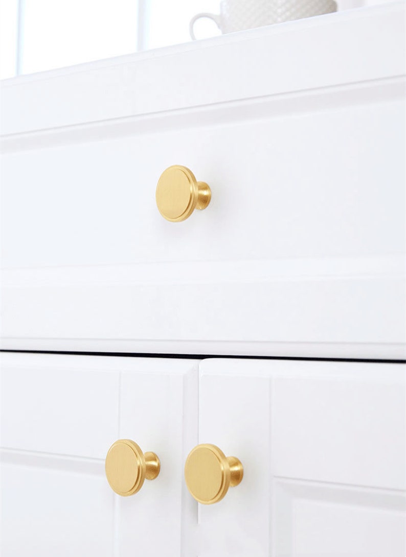 May include: Close-up of white cabinet doors and drawer with gold-colored, round cabinet knobs. The knobs have a flat, disc-shaped face and a short, cylindrical base. The cabinet doors and drawer are clean and modern.