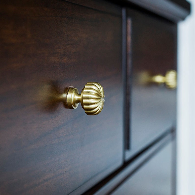 May include: Close-up of a dark brown wooden chest of drawers. The drawers have decorative, gold-coloured knobs with a ribbed, spherical design. The knobs are centered on the drawer fronts, adding a touch of elegance.