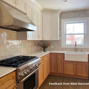 Puede incluir: Una cocina moderna con armarios superiores blancos e inferiores de madera natural. La cocina cuenta con una campana extractora de acero inoxidable, una cocina de gas y un fregadero blanco de granja. El salpicadero es de azulejos claros.