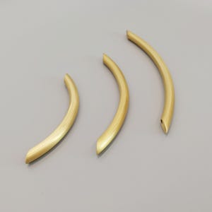 Peut inclure: Trois poignées de meuble en métal doré en forme de demi-cercle. Les poignées sont disposées en rangée, la plus grande à gauche et la plus petite à droite.