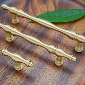 Peut inclure: Trois boutons de cabinet dorés avec un design décoratif. Les boutons sont disposés en rangée sur une surface en bois.