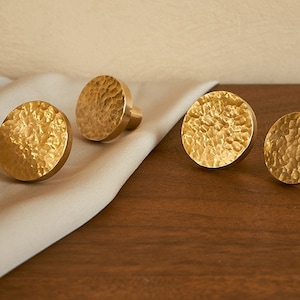 May include: Four gold-toned round cabinet knobs with a hammered texture. The knobs are arranged on a light brown surface and a beige fabric.