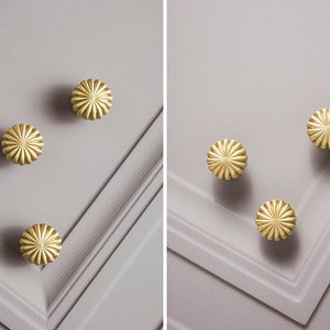 May include: Close-up of several gold-coloured, flower-shaped cabinet knobs. The knobs are arranged on a light grey surface with a textured, paneled design. The knobs have a detailed, ridged pattern.